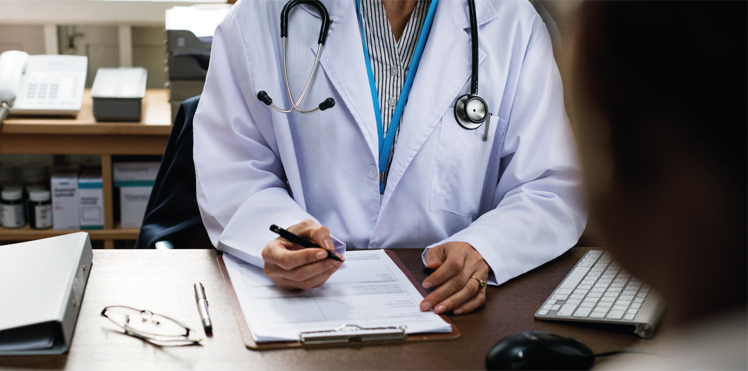 Profesional con túnica de médico blanca, estetoscópio colgado en el cuello. Está en su consultorio. Se ve un teléfono a lo lejos sobre una mesa de madera y unos estantes con medicamentos. En el esritorio de madera más oscura, al frente de la fotografía, hay un mouse negro, teclado blanco, lentes, carpeta, lapicera y planilla. Está entrevistando a un paciente, llenando la planilla con una lapicera. 